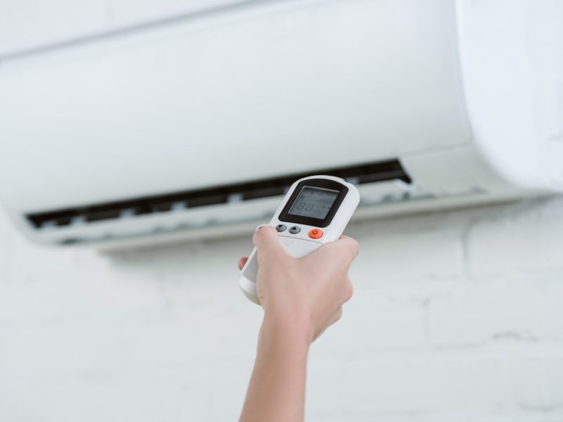 cropped shot of woman pointing at air conditioner hanging on brick wall with remote control